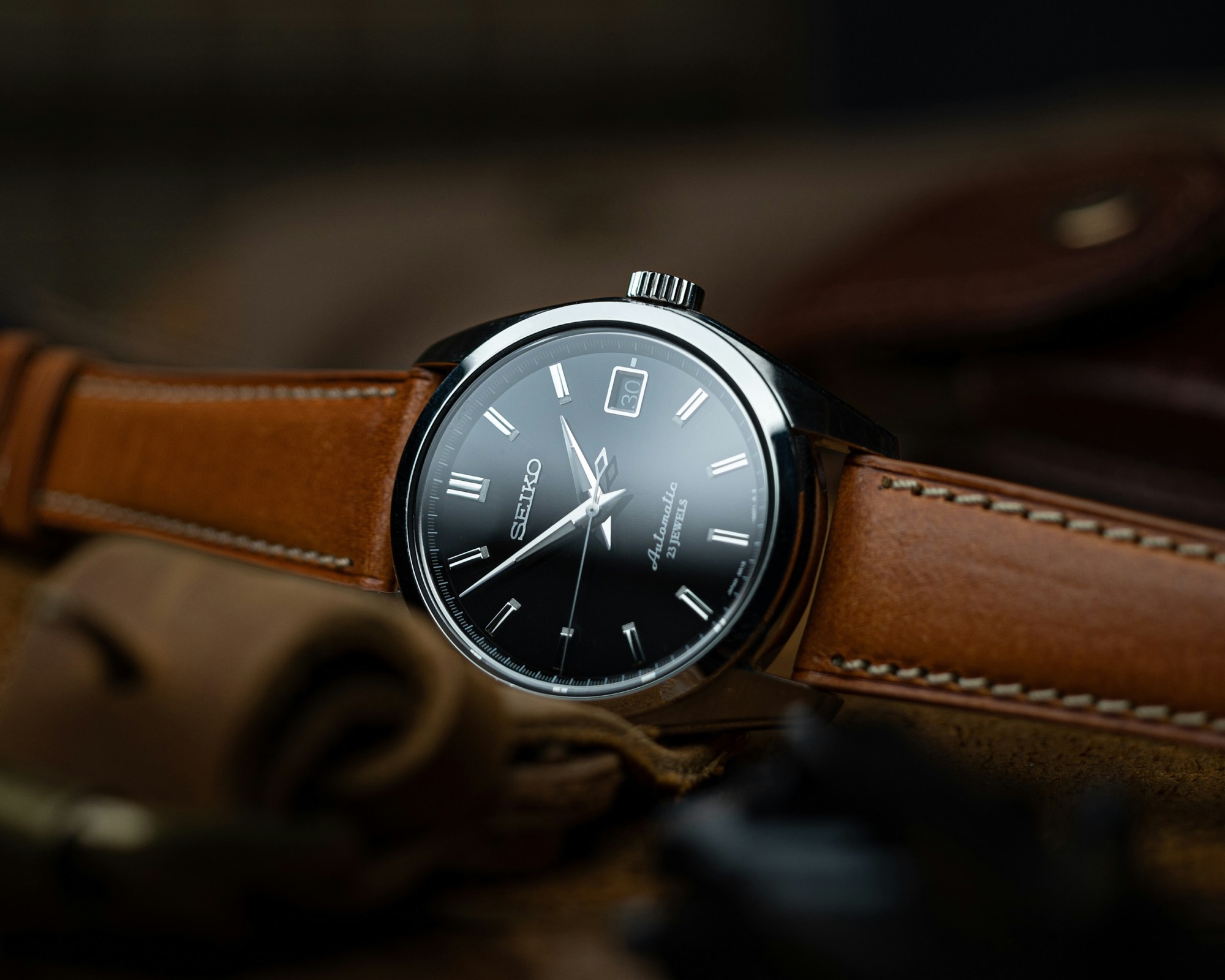 A black dial automatic watch on a tan brown leather strap in warm low light