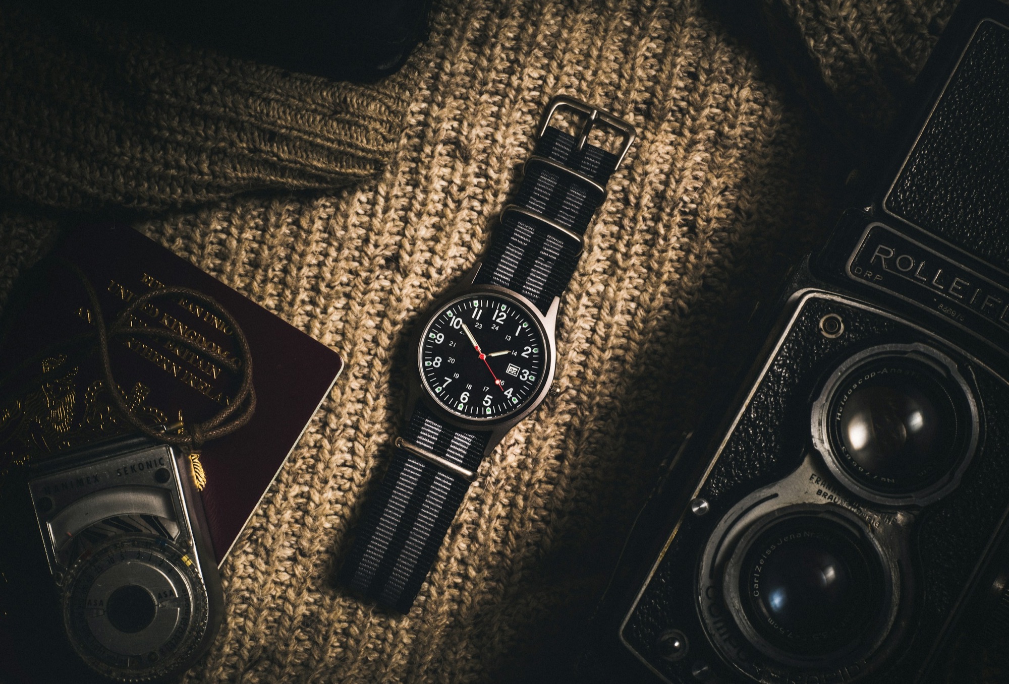 A field watch on a woven striped NATO strap laid on a knit fabric next to a vintage camera