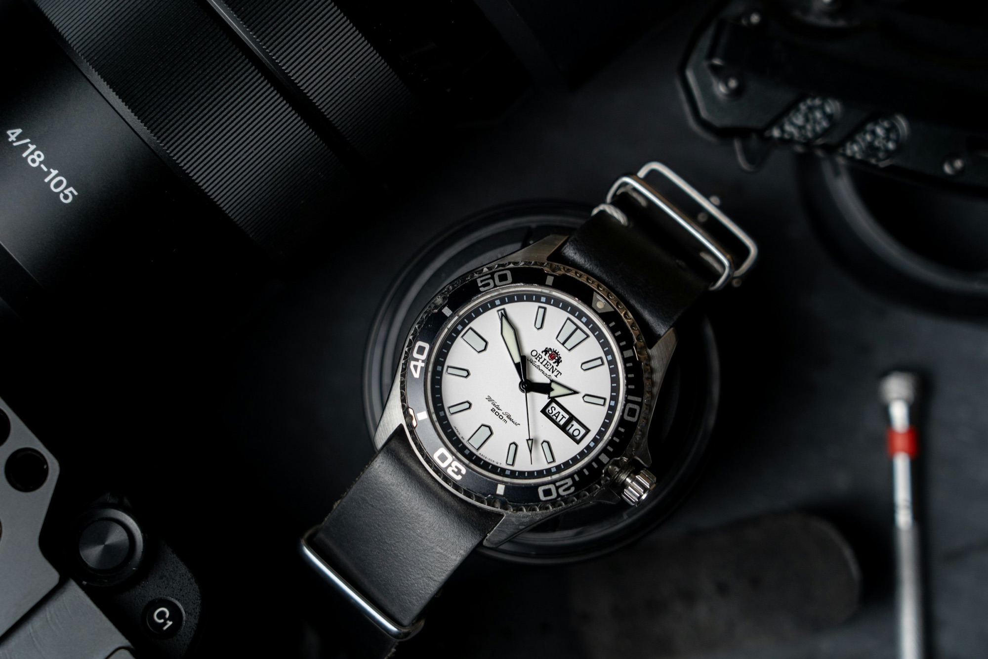 A white dial dive watch on a black NATO strap on a dark surface beside a camera and tools