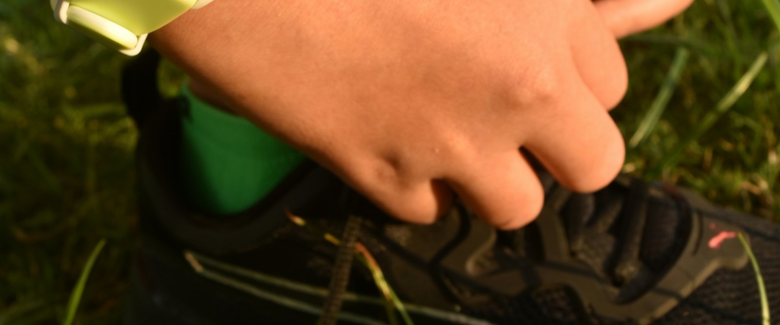 A sports watch on a pale rubber strap worn on the wrist while tying a running shoe on grass