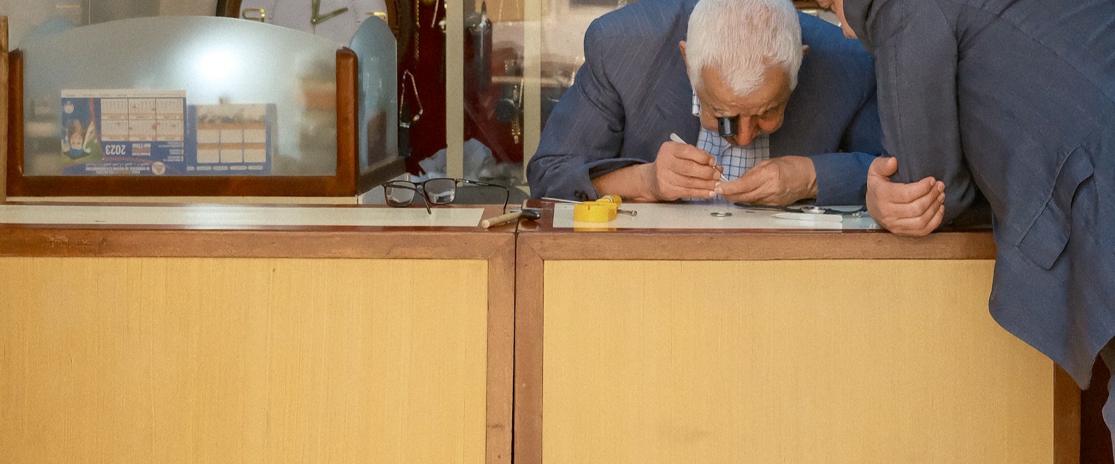 Two watchmakers leaning over a workshop counter examining a small timepiece, wall of clocks behind them