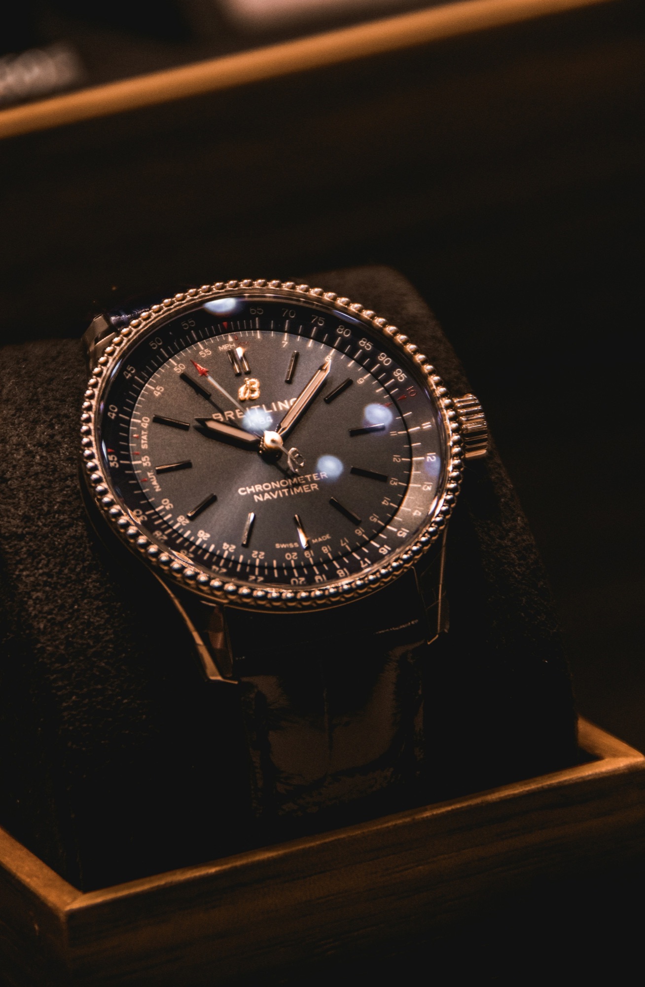 A dark dial sport chronograph watch displayed in a wooden case under moody light