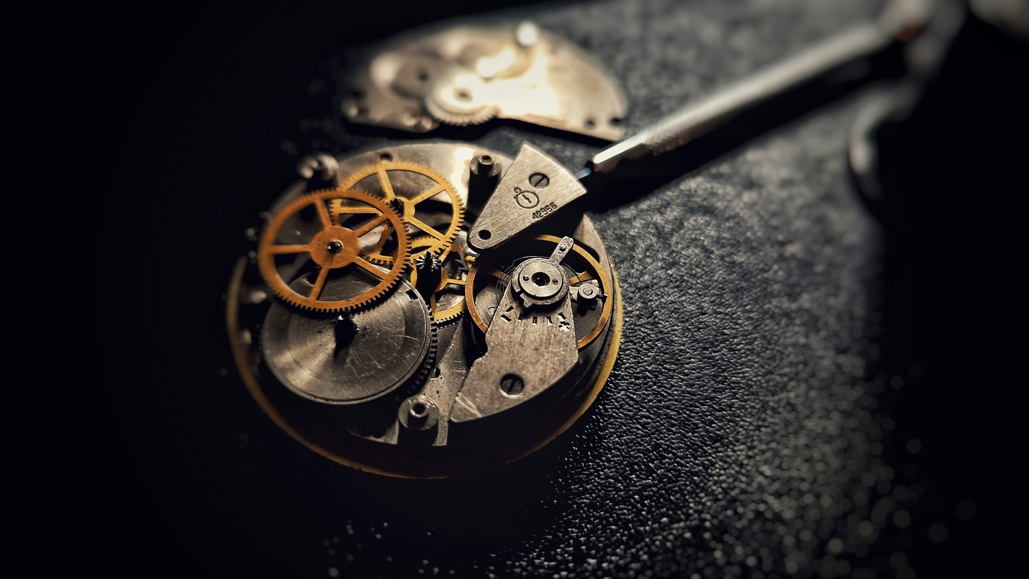 A close up of a watch movement with brass gears and a watchmaker screwdriver on a dark bench